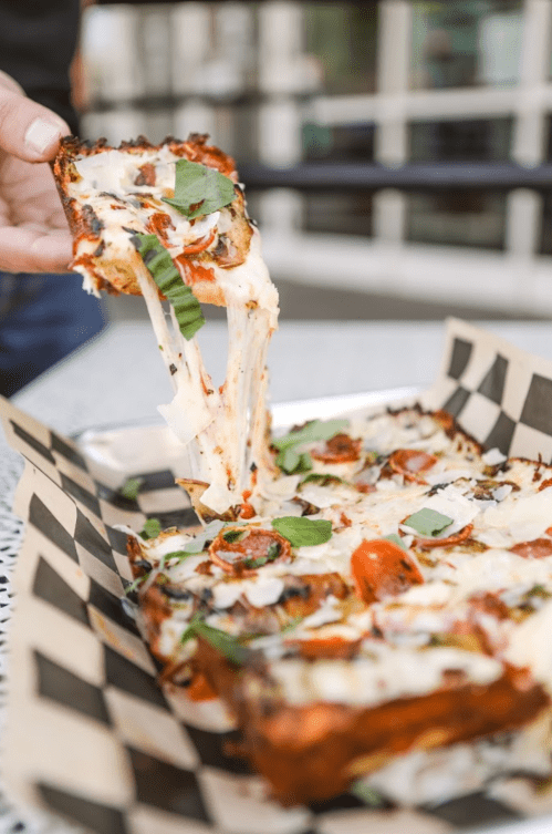 A hand lifts a slice of cheesy pizza topped with tomatoes and basil from a tray on a checkered paper.